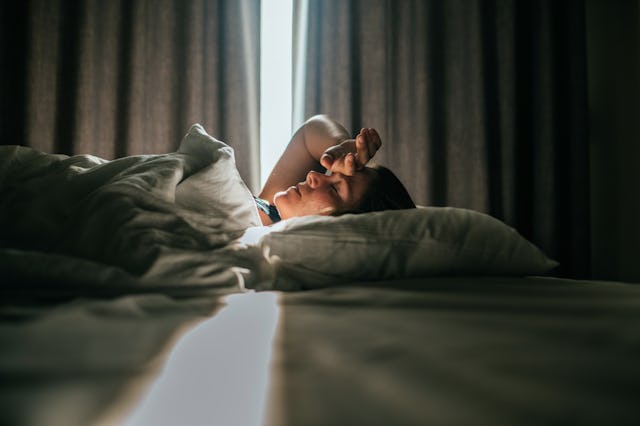 A person lies in bed, covered with a blanket, appearing to wake up. Sunlight streams through the par...